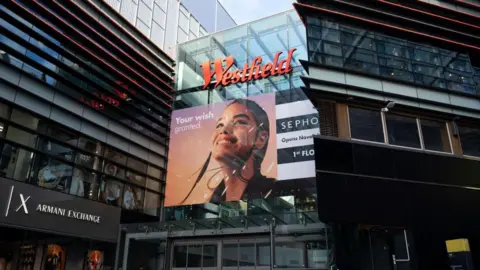 The outside of a large shopping centre with a glass and metal frontage. There is a large advert featuring the face of a young woman about the red Westfield sign above it. 