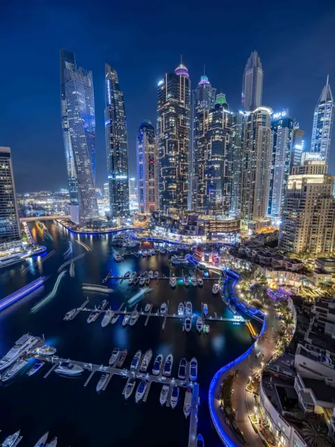 NurPhoto/Getty Images Dubai marina, tall illuminated glass buildings tower over a marina with a variety of boats. The image is taken at night.