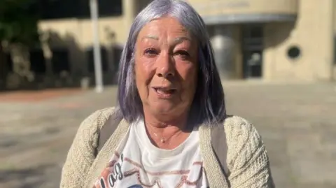 Emma Glasbey/BBC Angela Radford, a woman with purple-silver hair which is shoulder-length, wearing a patterned white t-shirt and cream cardigan standing outside court. 