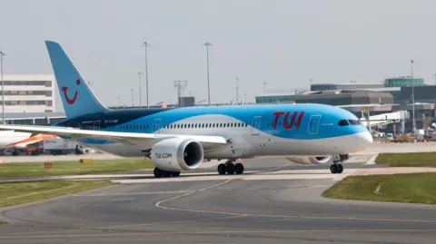 A blue and white Boeing 787 aircraft on the tarmac at Manchester Airport with terminal buildings behind it 