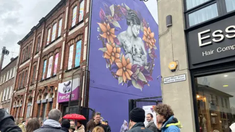 Picture of mural with purple background. It shows a mum breastfeeding a baby - part of which is censored. Around that is a reef with orange and purple flowers. 

Crowd of people are also in the picture with a pram centre and Great James Street sign. 