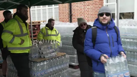 Three men in yellow hi-vis jackets standing behind slabs of bottled water. A man in a cobalt-coloured coat, wearing sunglasses, is holding a slab of bottled water. There is a woman, wearing a beige-coloured beanie, sunglasses and long black puffer jacket, also collecting bottled water. 