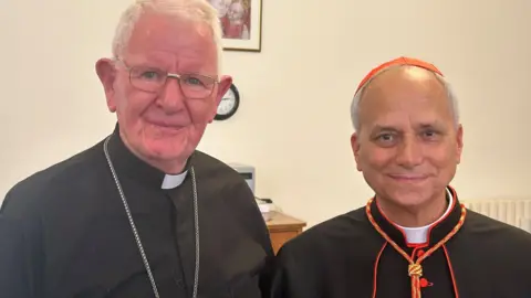 Bishop Michael Campbell Bishop Campbell (left) has grey hair, metallic spectacles and a silver chain and is wearing a white clerical dog collar with a black shirt. Bishop Robert Prevost, now Pope Leo XIV is wearing a black cardinal's soutane with red edgings and buttons, and a red skull cap. He has thin greying hair.