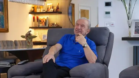 Lord Sugar wears a blue polo shirt and sits in a grey armchair