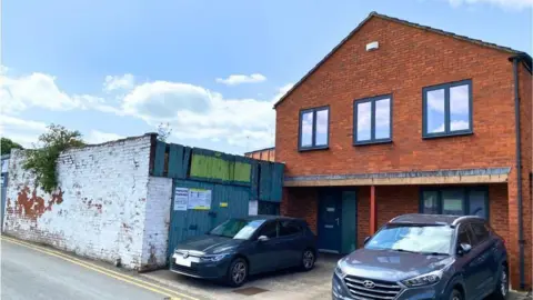 RRA Architects A red brick building with 9 windows. Two cars are parked in front. A wall with white paint can be seen on one side.