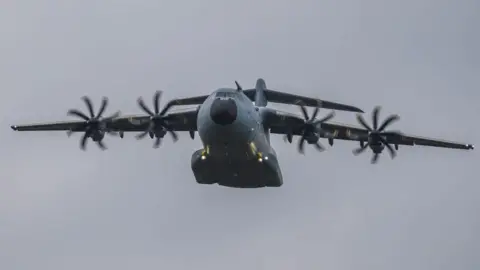 Dimitri Kuznecovas An Atlas A400M aircraft. The plane has four propellers and a broad grey body and raised tail and is in flight in the picture with the wheels raised up 