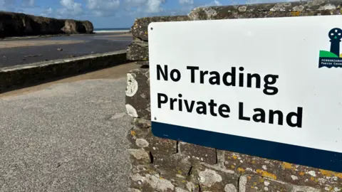 There is a metal sign in the right hand foreground reading No Trading Private Land, with Perranporth Parish Council logo beside it. it is on the edge of the approach to the beach with aa headland and the sea visible in the top left corner and a sandy beach beside it.