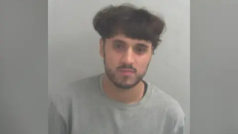 Essex Police Police custody photo of Ismaeel Malik, who is dressed in a grey prison-issue round-neck jumper. He has a short dark beard and has long hair on the top of his head, and short hair around the sides