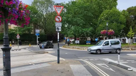 The Plain Roundabout in Oxford. In the foreground is the extremely sharp left turn from the Cowley Road to the Iffley Road. On the corner is a lamppost with a sign saying no left turns and an image of a camera. There are a few cars driving around. It's a clear day.