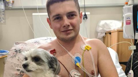 Louis in a hospital bed with wires attached to his chest, holding his beloved pet dog