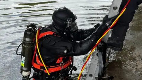 A diver in full black diving gear and breathing apparatus emerges from a canal and grips a metal ladder at the water’s edge. Bright coloured cables run from the equipment as another figure stands above, assisting from the bank.