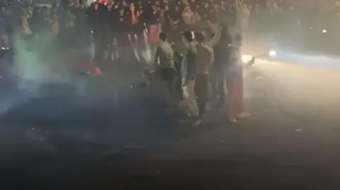 Two cars with headlights on driving around a circle of people at night. You can see the cars' lights beaming into the crowd of people.