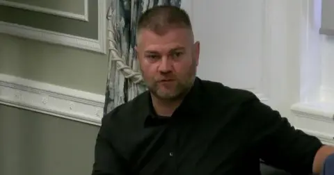 Colin McGrath has short cropped dark hair and a greying beard. He is wearing a dark shirt rolled up as the sleeves.