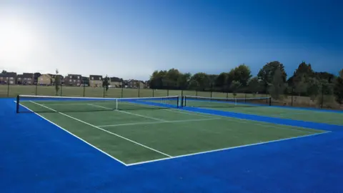 Peterborough City Council Two hard tennis courts with revamped surfaces. The courts are dark green and have a royal blue perimeter. Outside of the fenced courts is a public park with grass, surrounded by trees and houses. It is a sunny day.