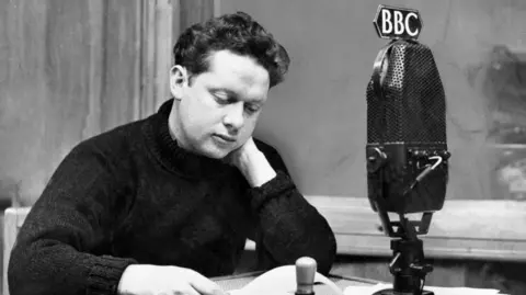 BBC Dylan Thomas sits at a table, the photograph is black and white. In front of him is a big microphone which has BBC on a sign. He appears to be reading something. 