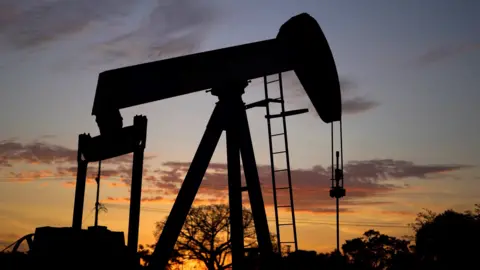 The silhouette of a nodding donkey oil pump against a twilight sky. The outline of a few trees can also be seen.