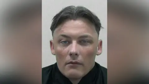 Mugshot of Morgan. He has thick dark and grey hair shaved at sides and long on top in a centre parting. He is clean shaven with a glazed expression.