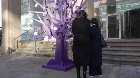 BBC Two women in longline coats stood lookig at a purple wooden tree figure with small cards attached to the braches.