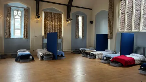 Hope Works A room, with Christmas lights put up on one wall, with nine beds, with bedding in them, and blue partitions. The floor is wooden, there are large curtains at a number of windows and the beds all have different bedding on. Boxes are at the end of the bed. 