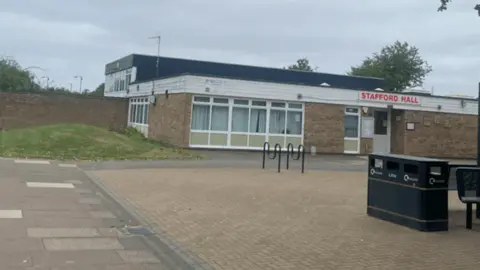 A redbrick modern building has large windows and white wooden cladding across the top. Above the door is a red sign saying Stafford Hall in upper case letters. There are bicycle racks outside and a bin and benches on a pave area in front.