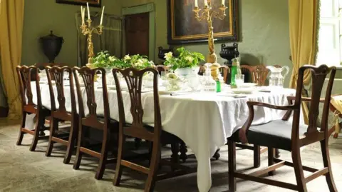 Rogers Jones andCo A long dining room table, draped with a white tablecloth and with green plants, gold candlesticks and expensive crockery on top of it. There are 10 wooden chairs around it, and in the background there are gold curtains and a painting on the wall.