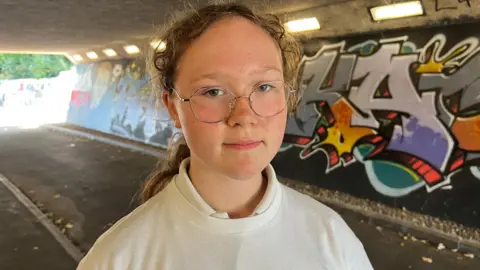 The picture shows 11-year-old Imogen standing in the underpass. She is wearing a white Citizens Somerset T-shirt. There is graffiti on the walls behind her.