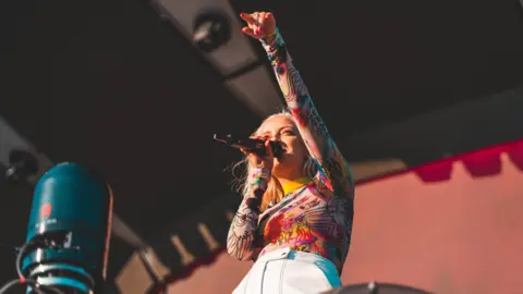Oli Hosier Singer Caswell in a colourful top with a microphone on a stage, with one arm in the air.