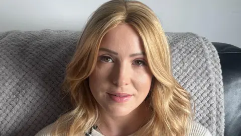 A woman with long blond hair looks into the camera. She is sitting on a sofa with a beige throw behind her. She is wearing a cream top.