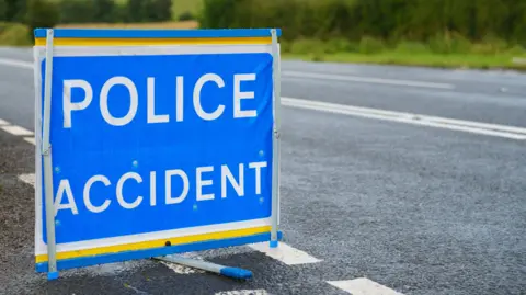 A square blue sign in the road which says 'Police Accident' on it in capital letters. There are no cars in view, only the tarmac with white road markings on it and greenery in the background along the side of the road. 