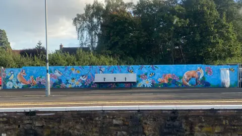 Chloe Farrant A wide shot of the mural which is painted on a long wall alongside the railway platform. A bench is in the middle of the wall, which is painted blue, with orange and white foxes, colourful birds, and flowers. 