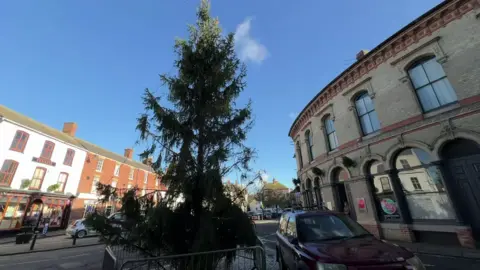 A market square with a Christmas tree, which appears to have several branches missing. The tree, which is reasonably tall,  is behind metal barriers.