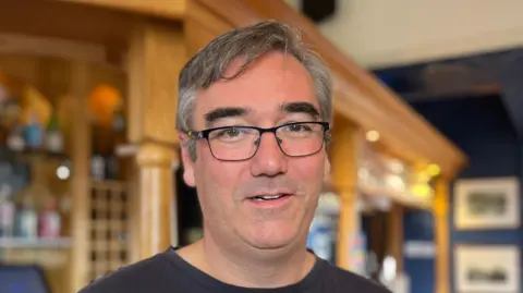 Tom Jackson/BBC Mark Whittington is looking straight at the camera. He is wearing glasses, a black T-shirt and is standing in a pub with the bar behind him.