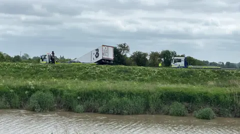 Quays Najm/BBC Several heavy goods vehicles are on a road. One of them is a lorry which has collided into bushes. In the foreground is a river which then leads to a grass verge.