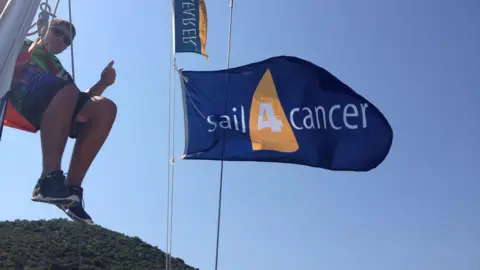 Matt Tozer A teenage boy sat in the riggings of a sail boa, with a flag of an organisation which gives sailing trips to children with cancer in the foreground.