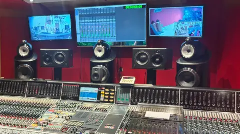 Three screens can be seen above a mixing desk with various speakers and recording instruments also in vision.