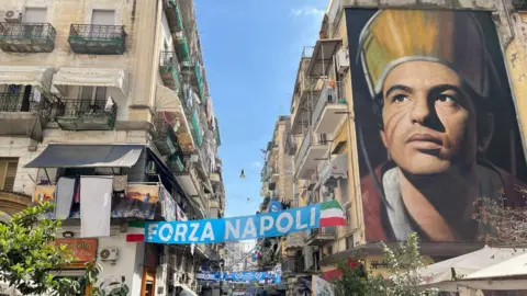 Huge wall mural of Scott McTominay in a Naples street. There are Napoli banners strung between flats on either side of the street