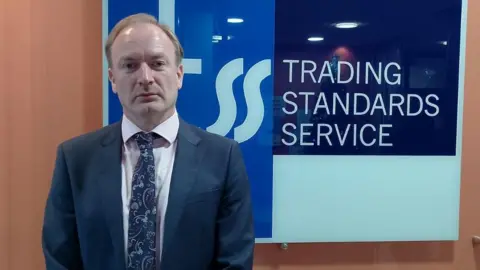 Damien Doherty looking to camera, standing in front of a blue and white Trading Standards Service sign.  He has short, fair hair and is wearing a navy suit, a navy patterned tie and a light-coloured shirt. 