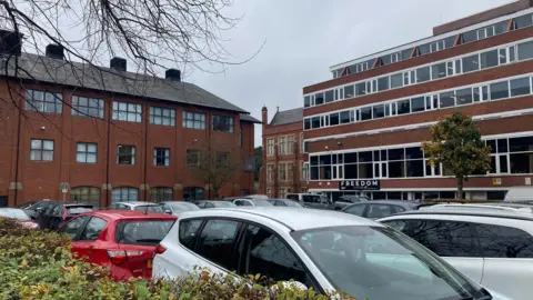 A number of cars parked in a parking lot. The car park is surrounded by large red brick buildings. 