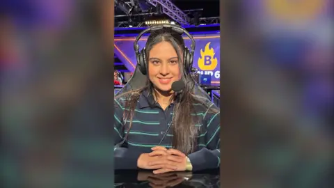 A young woman is sitting in a computer chair, in an e-sport venue. She is looking directly into the camera, smiling. She is wearing a striped shirt and a headset.