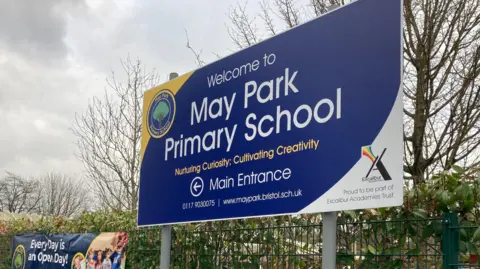 The exterior entrance of the school. Outside there is a blue sign, some bushes, and blue bollards shaped to look like pencils.