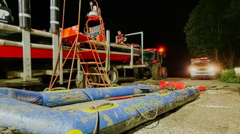 Humber Rescue A long blue inflatable raft is lying in the foreground covered in mud with "Humber Rescue" on it. In the background a tractor is pulling another Humber Rescue craft along the riverside. The scene is illuminated and another rescue vehicle has its lights on.