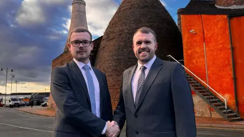 Reform UK Luke Shenton and Jonathan Gullis (right). Both are wearing dark suits and smiling at the camera as they shake hands. in the background is a traditional Stoke-on-Trent ceramics industry kiln