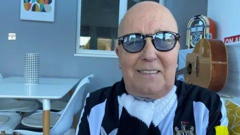 BBC Paul Atkinson is a 69-year-old man sitting in his house wearing a black and white scarf and top. Behind him is a guitar, a small juke box, an "on air" sign and a white dining table with four white chairs. He is wearing glasses and is bald 