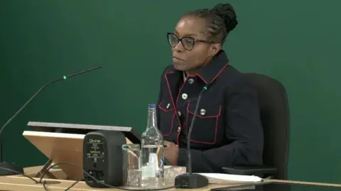 A woman sat a desk in a blue jacket with red trim in front of a green back drop.