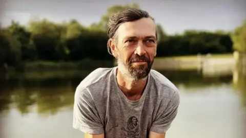 Mr Thompson is wearing a grey T-shirt and is sat in front of a lake. He has a beard and brown hair. He is looking slightly past the camera.