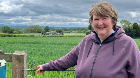 A woman in a purple top by a field gate with a military helicopter in the distance