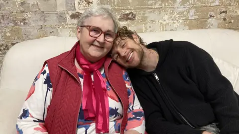 BBC Tom Grennan, who is wearing a black hoodie, leaning his head on his gran's shoulder. She has short grey hair with red glasses, and a red jacket with red scarf.