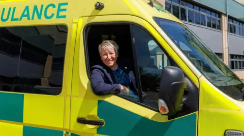 MP Rachel Taylor is sitting at the wheel of an ambulance which is liveried in yellow and green. It is parked outside an office building.