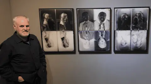 Paul Ford has his hands in his pockets and is standing by three frames of black-and-white blended photographs. One is of a young girl, one is of an old man, and one is of two young children (Ford and his sister).