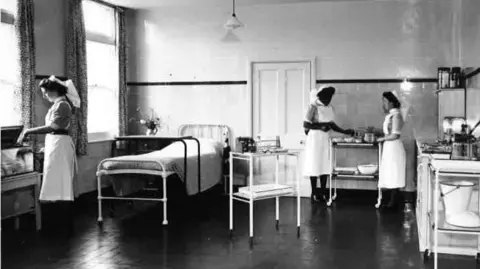 South Warwickshire University NHS Foundation Trust Black and white photo featuring a metal hospital bed. Two nurses wearing white aprons and caps stand by a trolley. A third stands next to what looks like an incubator.